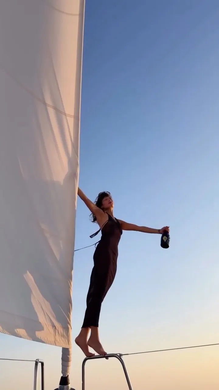 Mimi standing on the bow of a sailing boat against a pastel evening sky