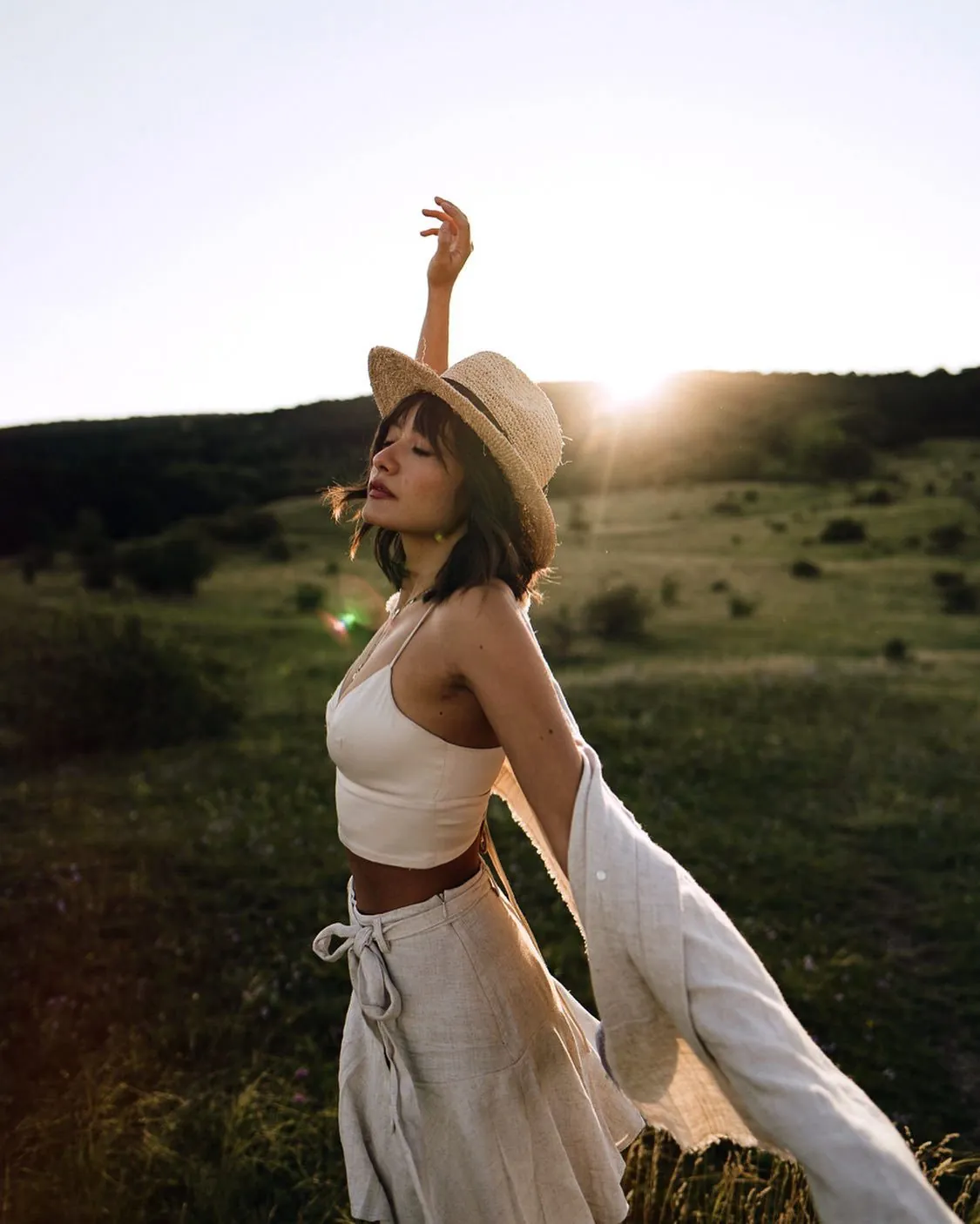 Mimi in a straw hat and linen, arms raised in golden-hour light on a meadow
